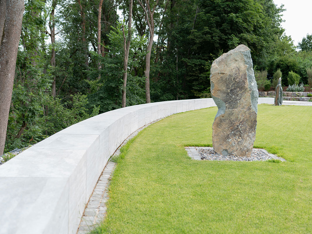 Grüner Rasen mit einer Steinskulptur, links davon eine niedrige Betonmauer.