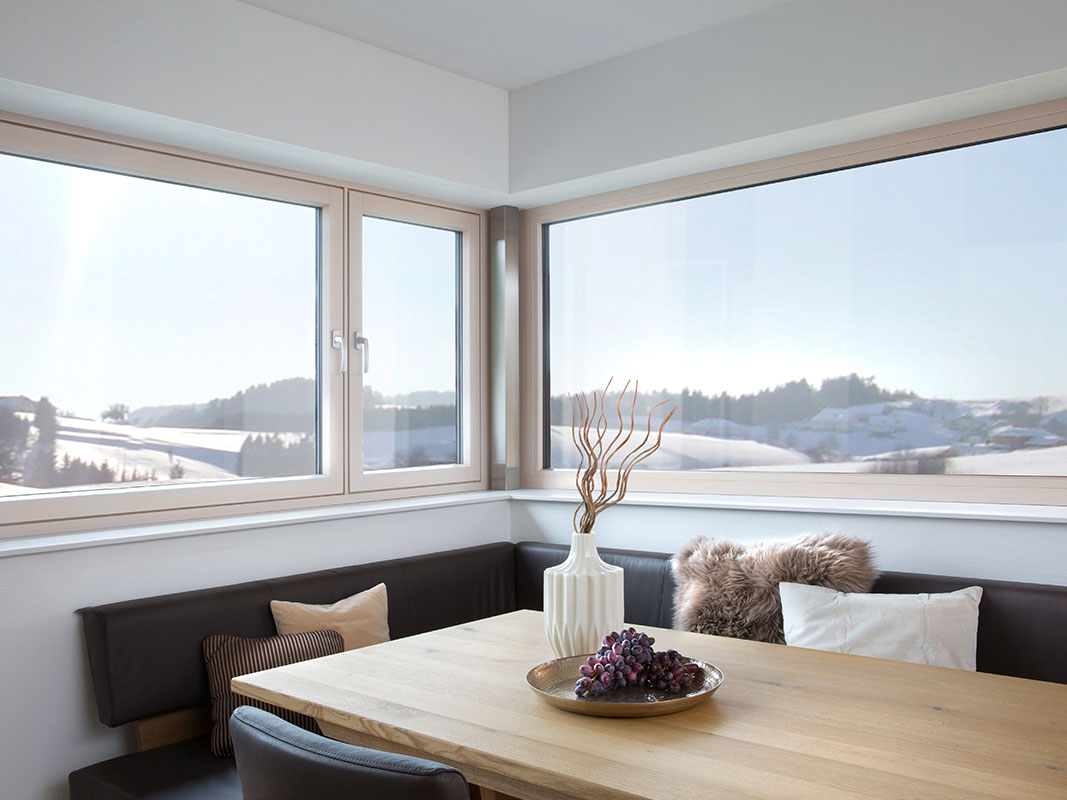 Sitzecke mit dunklem Ledersofa, Holztisch und Plüschkissen vor einer großen Fensterfront, dahinter eine verschneite Winterlandschaft.