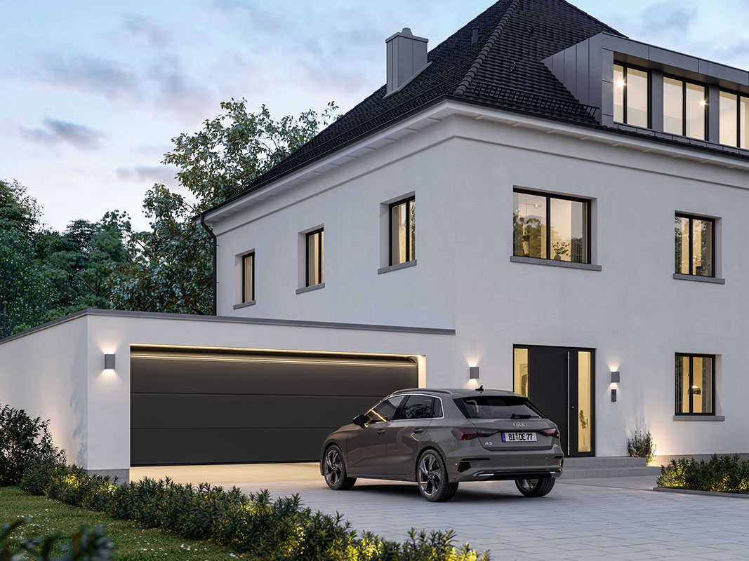 Großes dunkles Sektionaltor einer Garage, davor ein Auto beim Einfahren, Lichtstrahl aus der Garage und beleuchtetes Haus im Hintergrund während der Dämmerung.