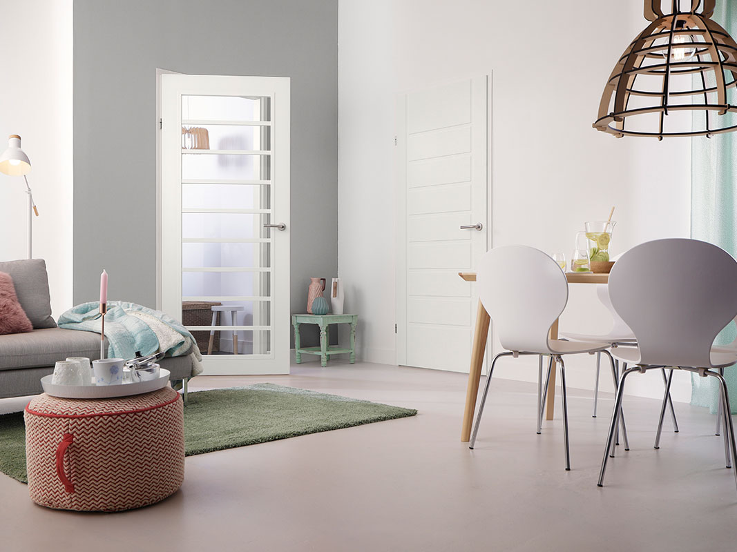 Wohnzimmerausschnitt mit verglaster Tür, Esstisch mit weißen Stühlen rechts und Sofa mit Decke links.