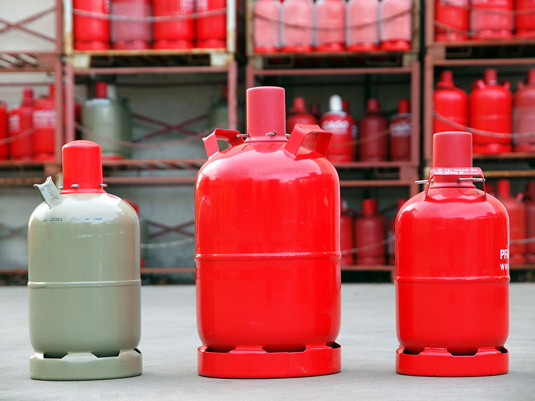 Drei Propangasflaschen vor einem Regal voller Flaschen: mittlere Flasche groß und rot, rechte Flasche klein und rot, linke Flasche klein und beige.