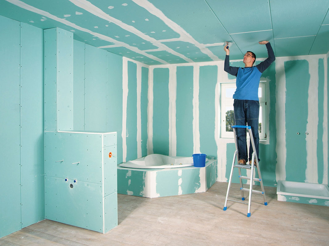 Handwerker auf einer Leiter verspachtelt blaue Dämmplatten in einem Rohbau-Raum.