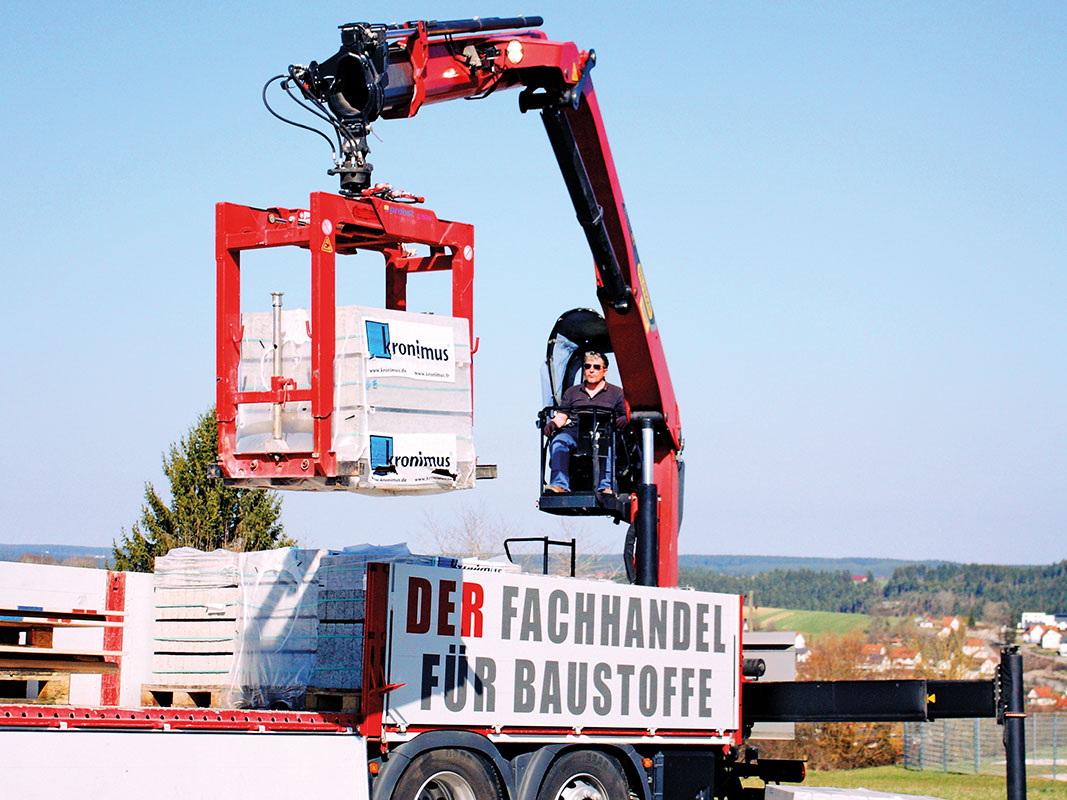 Ein Mann sitzt in einem roten Kran, der Paletten mit Baustoffen für den Hausbau nach oben hebt.
