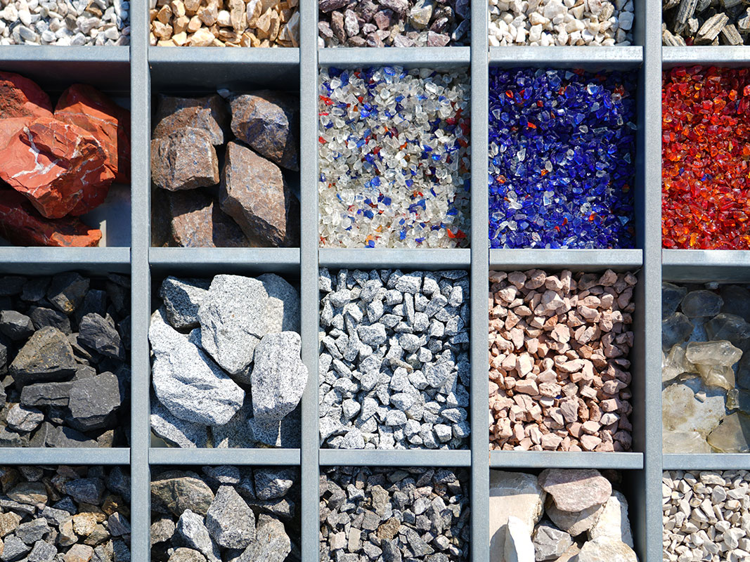 Von oben fotografierte Schublade mit mehreren Fächern, die verschiedene Steinsorten enthalten – von großen schwarzen Steinen bis zu blauen und roten Glassteinen.